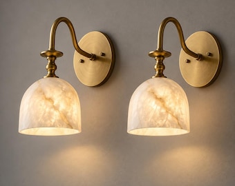 May include: A pair of wall sconces featuring curved brass arms and circular wall plates. Each fixture has a dome-shaped shade made of translucent white stone with natural veining, emitting a warm glow from the interior light source.