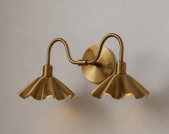 May include: A double-arm wall sconce finished in brushed brass. The fixture features a circular wall plate and two curved arms extending outward, each supporting a downward-facing, fluted, bell-shaped shade with a scalloped edge design.