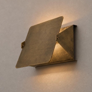May include: A wall-mounted light fixture crafted from aged brass with a geometric, angular design. The fixture features a large, flat, square-shaped front plate that partially overlaps a triangular side panel, casting a warm glow from within.