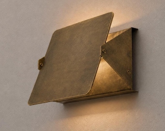 May include: A wall-mounted light fixture crafted from aged brass with a geometric, angular design. The fixture features a large, flat, square-shaped front plate that partially overlaps a triangular side panel, casting a warm glow from within.