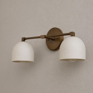 May include: A wall-mounted light fixture featuring a circular antique brass backplate and a horizontal arm supporting two downward-facing, dome-shaped matte white glass shades. The metal hardware has a warm, metallic finish throughout.