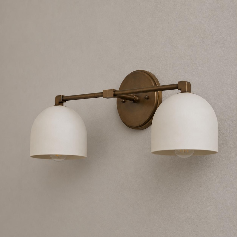 May include: A wall-mounted light fixture featuring a circular antique brass backplate and a horizontal arm supporting two downward-facing, dome-shaped matte white glass shades. The metal hardware has a warm, metallic finish throughout.
