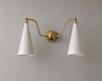 May include: A double wall sconce featuring two white cone-shaped shades attached to a central brass circular mounting plate by two curved brass arms. The fixture has a polished brass finish on the hardware and matte white metal conical shades.