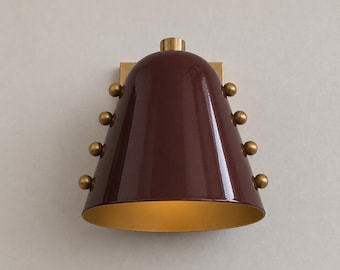 May include: A wall sconce featuring a glossy, deep burgundy bell-shaped shade with a gold interior. The exterior of the shade is accented with four small, round, brass-toned spheres on each side, and it is mounted on a square brass backplate.