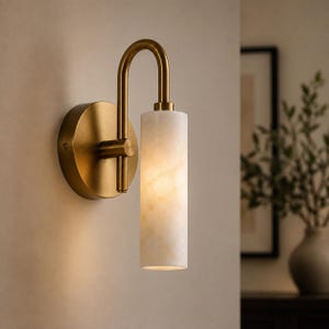 May include: A wall sconce featuring a circular brass backplate and a curved arm that supports a cylindrical, translucent white stone shade. The fixture emits a warm, soft glow from within the stone, highlighting its natural veined texture.
