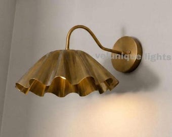 May include: A wall-mounted lamp featuring a brass-toned, fluted, bell-shaped shade with a scalloped edge. The fixture includes a curved arm extending from a circular wall plate. The text "velunique lights" is visible on the wall near the lamp.