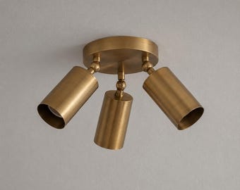 May include: A three-light ceiling spotlight fixture featuring a circular brass base and three adjustable cylindrical brass heads. Each light head is connected to the central mount by a swivel joint, allowing for directional lighting adjustments.