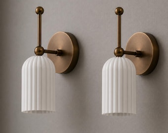 May include: A pair of wall sconces featuring a circular bronze-toned backplate, a vertical rod with a spherical finial, and a white, vertically ribbed cylindrical shade. The metal components have a smooth, matte finish, while the shade is opaque.