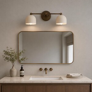 May include: A two-light wall sconce with a dark bronze finish and matte cream dome shades mounted above a rectangular mirror with a thin bronze frame. The fixture features a horizontal bar design and emits a warm glow from both light bulbs.