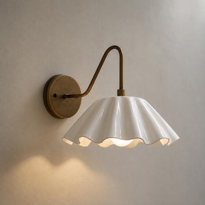 May include: A wall-mounted light fixture featuring a circular antique brass backplate and a curved arm extending to a white, glossy, scalloped ceramic shade. The shade has a wide, fluted edge and houses a single light bulb emitting a warm glow.