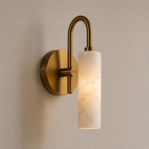 May include: A wall sconce featuring a circular brass mounting plate and a curved arm that supports a vertical, cylindrical white stone shade with a marbled texture. The fixture has a metallic finish and emits a soft, warm glow from the stone.