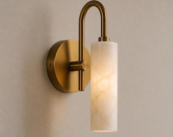 May include: A wall sconce featuring a circular brass mounting plate and a curved arm that supports a vertical, cylindrical white stone shade with a marbled texture. The fixture has a metallic finish and emits a soft, warm glow from the stone.