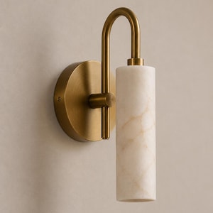 May include: A wall sconce featuring a circular brushed brass backplate and a curved arm supporting a cylindrical white marble shade with subtle beige veining. The fixture has a vertical orientation with the light source directed downward.