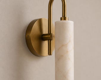 May include: A wall sconce featuring a brushed brass finish circular backplate and a curved arm supporting a cylindrical white marble shade with subtle beige veining. The fixture has a vertical orientation with the light opening facing downward.