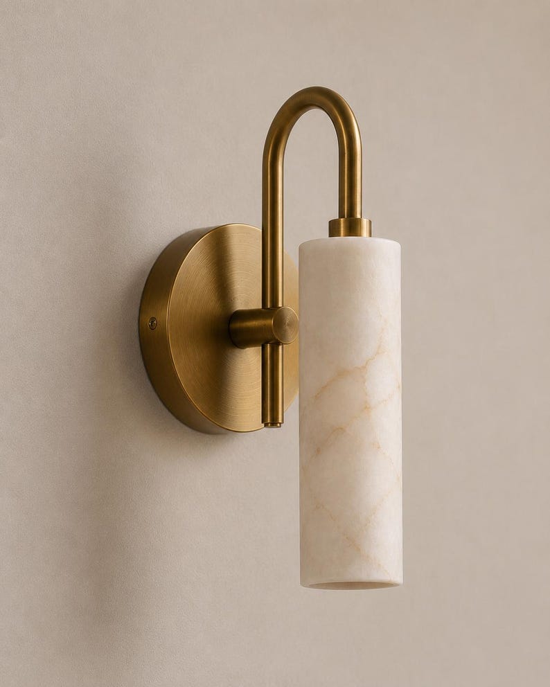 May include: A wall sconce featuring a circular brushed brass backplate and a curved arm supporting a cylindrical white marble shade with subtle beige veining. The fixture has a vertical orientation with the light source directed downward.