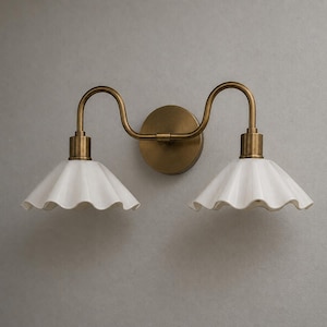 May include: A double wall sconce featuring a circular brass backplate and two curved brass arms extending outward. Each arm supports a white, fluted, bell-shaped shade with a scalloped edge, creating a symmetrical lighting fixture design.