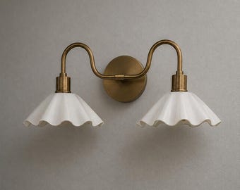 May include: A double wall sconce featuring a circular brass backplate and two curved brass arms extending outward. Each arm supports a white, fluted, bell-shaped shade with a scalloped edge, creating a symmetrical lighting fixture design.