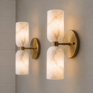May include: Two wall sconces featuring dual cylindrical white marble shades with grey veining. Each fixture is mounted on a circular brushed brass backplate with a central horizontal arm connecting the two stacked stone light diffusers.