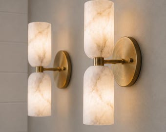 May include: Two wall sconces featuring dual cylindrical white marble shades with grey veining. Each fixture is mounted on a circular brushed brass backplate with a central horizontal arm connecting the two stacked stone light diffusers.