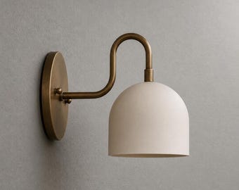 May include: A wall-mounted light fixture featuring an oval-shaped antique brass backplate and a curved arm that supports a matte white dome-shaped shade. The metal arm connects to the shade with a cylindrical fitting, directing light downward.