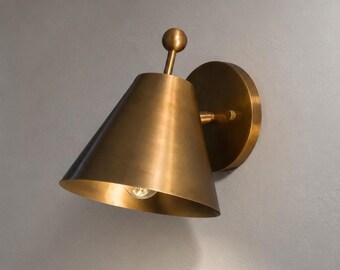 May include: A wall-mounted lamp with a conical brass shade and a circular backplate. The fixture features a small vertical stem topped with a spherical finial extending from the top of the cone, with a single light bulb visible inside the shade.