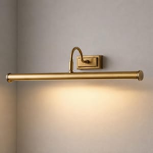 May include: A wall-mounted picture light with a brushed brass finish. The fixture features a rectangular base plate, a curved arm, and a long, horizontal cylindrical lamp head that casts a warm, downward-facing glow along the wall surface.