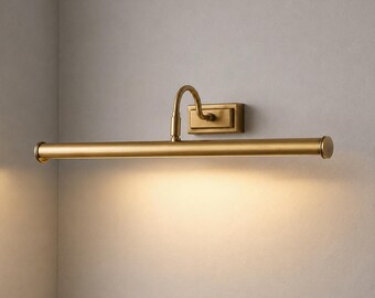 May include: A wall-mounted picture light with a brushed brass finish. The fixture features a rectangular base plate, a curved arm, and a long, horizontal cylindrical lamp head that casts a warm, downward-facing glow along the wall surface.
