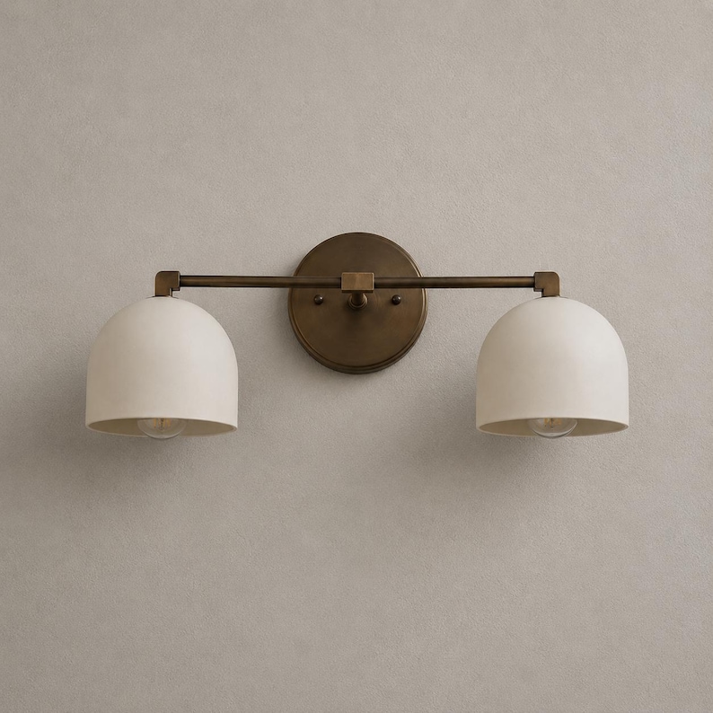 May include: A two-light wall sconce featuring a circular antique brass backplate and a horizontal bar supporting two matte off-white dome-shaped shades. The fixture has a metallic finish on the central mounting plate and the connecting hardware.