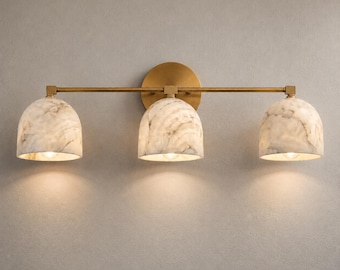 May include: A three-light wall fixture featuring a horizontal brass bar and a circular brass wall mount. Three dome-shaped shades made of white and grey marbled stone are attached to the bar, each housing a single light bulb pointing downward.