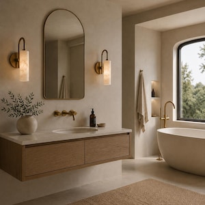 May include: A bathroom vanity with a light wood fluted drawer front, a white stone countertop, and a round-top mirror. Two cylindrical wall sconces with warm light flank the mirror, accompanied by a brass faucet and a ceramic vase with greenery.