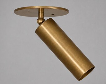 May include: A cylindrical brass spotlight fixture featuring a circular mounting plate and an adjustable swivel joint. The metal surface has a matte finish, and the design includes a hollow tube structure that is angled downward from the base.
