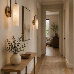 May include: Two wall-mounted light fixtures featuring curved brass arms and cylindrical white marble shades with subtle veining. The lamps emit a warm glow, illuminating a hallway with a wooden console table, a ceramic vase, and a bowl.