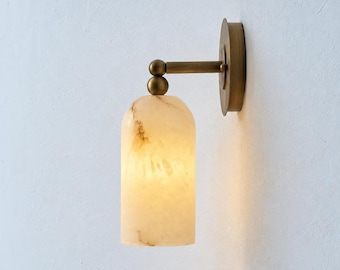 May include: A wall-mounted light fixture featuring a cylindrical, cream-colored shade with brown marble-like veining. The fixture includes a brass-toned metal arm and a circular wall plate, connected by two spherical brass-toned joints.