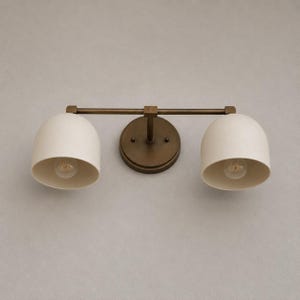 May include: A two-light wall sconce featuring a dark bronze finish metal frame and two dome-shaped off-white shades. The fixture includes a circular wall mounting plate connected to a horizontal bar that supports the two downward-facing light bulbs.