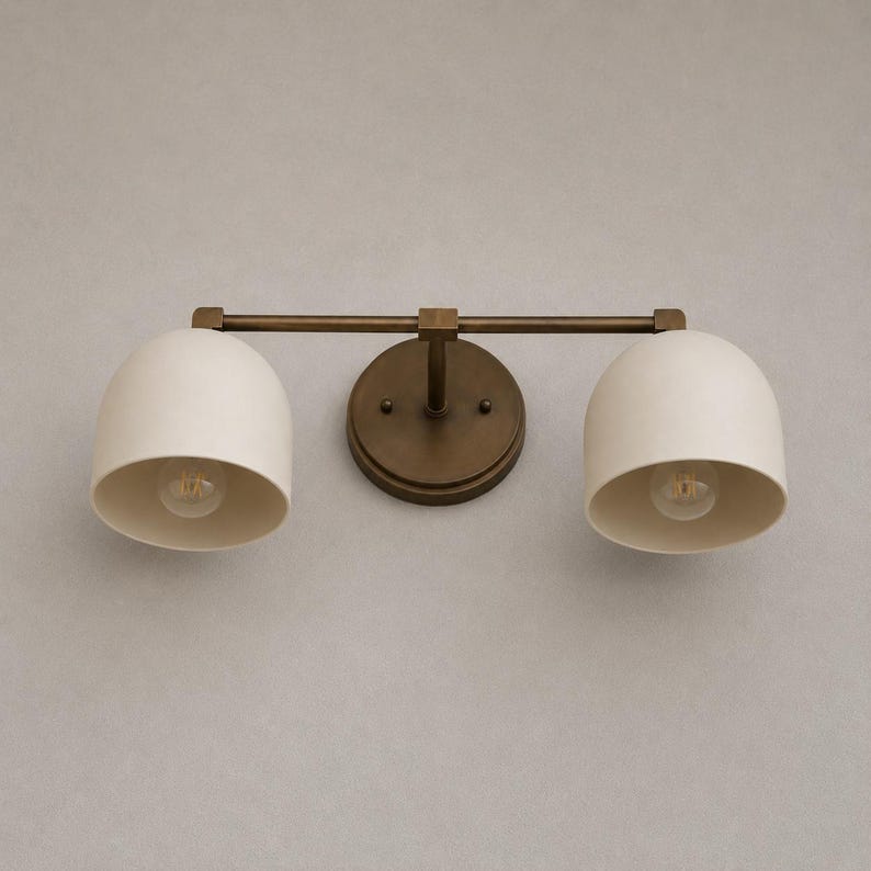 May include: A two-light wall sconce featuring a dark bronze finish metal frame and two dome-shaped off-white shades. The fixture includes a circular wall mounting plate connected to a horizontal bar that supports the two downward-facing light bulbs.