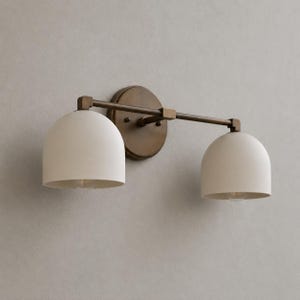 May include: A two-light wall sconce featuring a dark bronze finish metal frame and two dome-shaped off-white matte shades. The fixture includes a circular wall mounting plate connected to a horizontal bar that supports the two downward-facing lights.