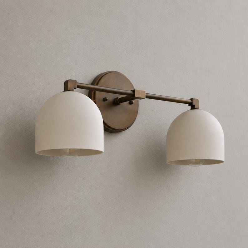May include: A two-light wall sconce featuring a dark bronze finish metal frame and two dome-shaped off-white matte shades. The fixture includes a circular wall mounting plate connected to a horizontal bar that supports the two downward-facing lights.