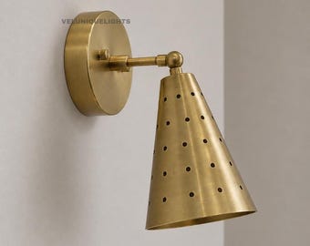 May include: A brass wall sconce featuring a circular mounting plate with the text VELUNIQUELIGHTS and a conical shade with a perforated dot pattern. The fixture includes an adjustable arm with a swivel joint connecting to the metal cone.