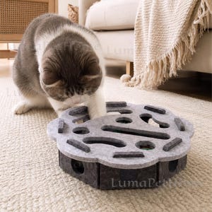 以下が含まれることがあります： グレーと黒の猫が、グレーのフェルト製の猫用おもちゃで遊んでいます。おもちゃには、インタラクティブな遊びができるように、複数の穴とスロットがあります。「LumaPetHome」の文字が見えます。