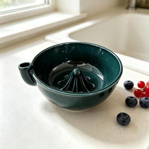 May include: A dark forest green ceramic citrus juicer featuring a central ribbed reamer and a small pouring spout on the side. The bowl has a smooth, glossy finish and is accompanied by a few scattered fresh blueberries and red currants.