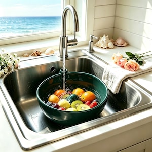 May include: A stainless steel sink with a green bowl filled with fresh fruit, including oranges, lemons, kiwi, and raspberries. A faucet pours water into the bowl. The scene is set near a window with a view of the ocean.