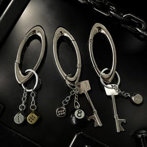 Puede incluir: Tres llaveros de mosquetón de metal plateado. El primero tiene dos dijes de dados. El segundo incluye un dije de dado, una bola de billar número ocho y una llave antigua. El tercero presenta una llave antigua y un dije de dado.