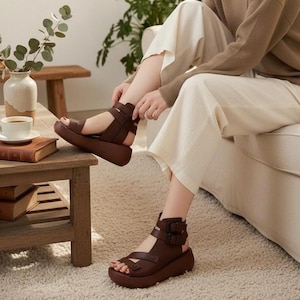 May include: A pair of dark brown leather platform sandals featuring an ankle strap with two adjustable buckles, a thick sole, and an open-toe design. The sandals are worn with light-colored wide-leg trousers and a long-sleeved brown sweater.