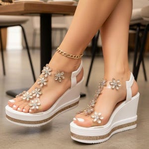 Könnte beinhalten: Weiße Plateau-Keilsandalen mit transparenten Riemen, die mit funkelnden Blumen verziert sind. Die Plattform und der Keilabsatz sind weiß, mit einem goldfarbenen Glitzersaum. Ein goldfarbenes Fußkettchen ist ebenfalls zu sehen.