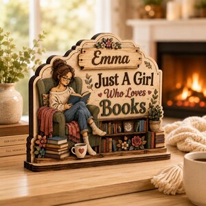 May include: A decorative wooden sign featuring a layered illustration of a person reading in a green armchair. The text reads Emma Just A Girl Who Loves Books. The design includes a bookshelf, stacks of books, a mug with a heart, and floral accents.