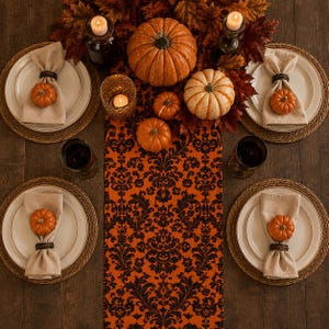 May include: A table setting featuring four white plates on woven chargers, each with a beige napkin held by a dark ring and a small orange pumpkin. A central orange runner with a black damask pattern holds pumpkins, candles, and autumn leaves.