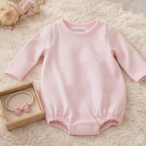 May include: A pink and white striped baby romper with long sleeves. A matching pink bow headband is displayed in a small wooden box. The romper and headband are on a soft, white, fluffy surface.