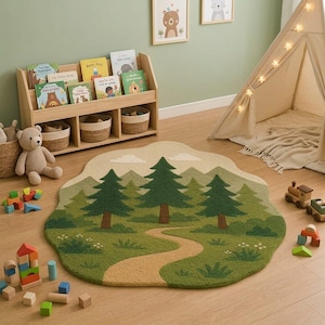 May include: A tufted area rug featuring a landscape design with green pine trees, a winding tan path, and light green mountains against a cream sky. The rug has an irregular, cloud-like shape and is placed on a wooden floor in a room with toys.
