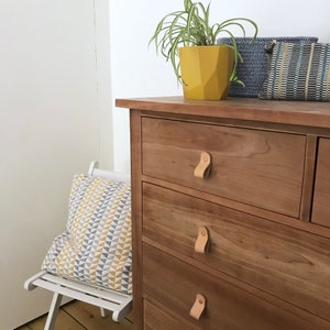 May include: A wooden chest of drawers with four drawers and leather pulls. The dresser is made of light wood and has a natural finish. A small plant in a yellow pot sits on top of the dresser.