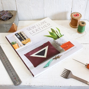 May include: A book titled "Sew Luxe Leather: Over 20 Stylish Leather Craft Accessories" by Rosanna Clare Gethin. The book cover features a burgundy leather pouch with a black and white triangle design.